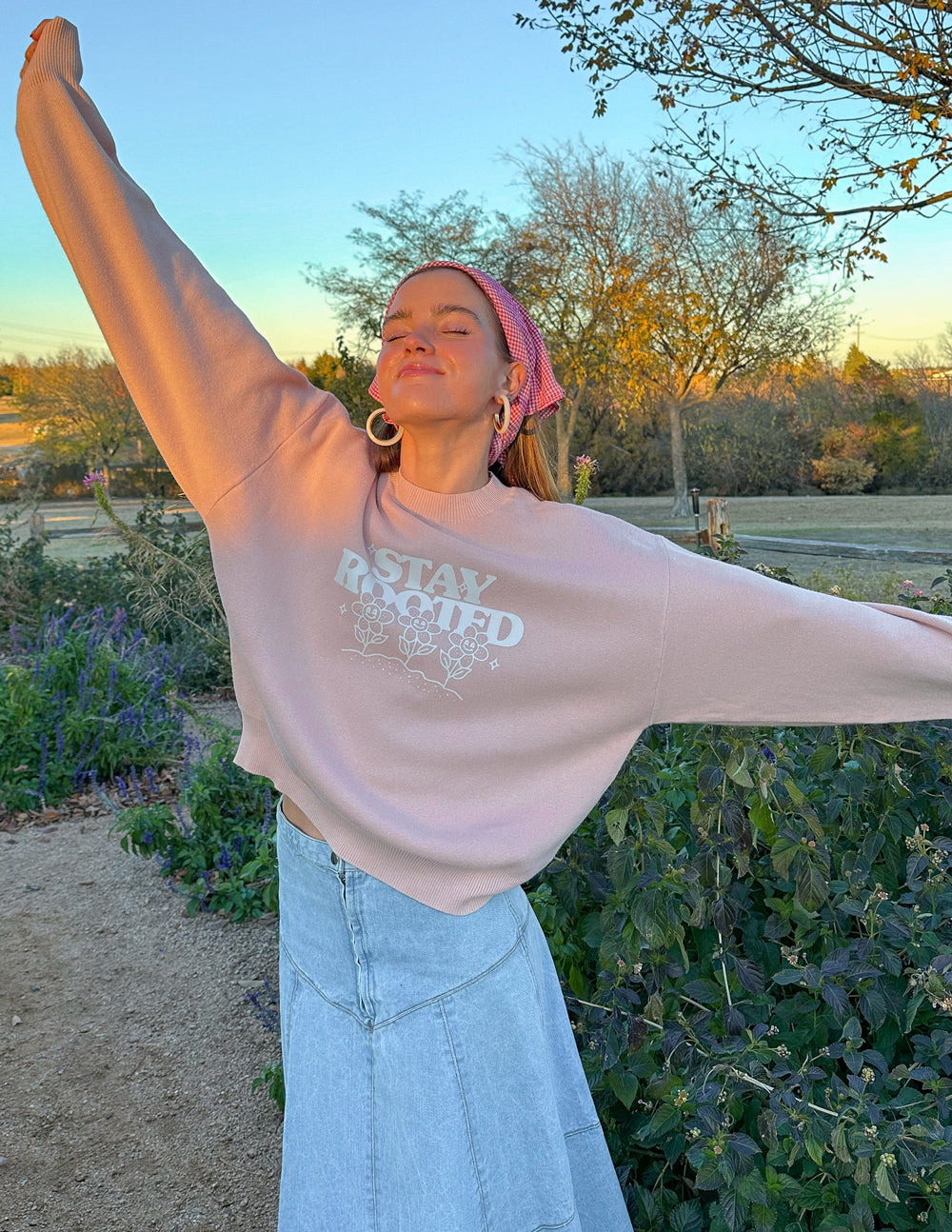 Person wearing a light pink sweatshirt with text and light blue jeans, standing outdoors with arms outstretched.
