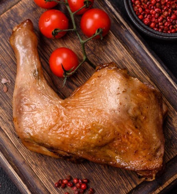 Roasted pasture-raised chicken leg on a rustic wooden cutting board with fresh cherry tomatoes and red peppercorns.
