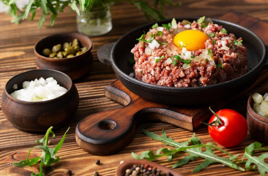 Raw premium Angus ground beef topped with a fresh egg yolk and diced onions in a cast iron skillet, surrounded by rustic ingredients on a wooden table.