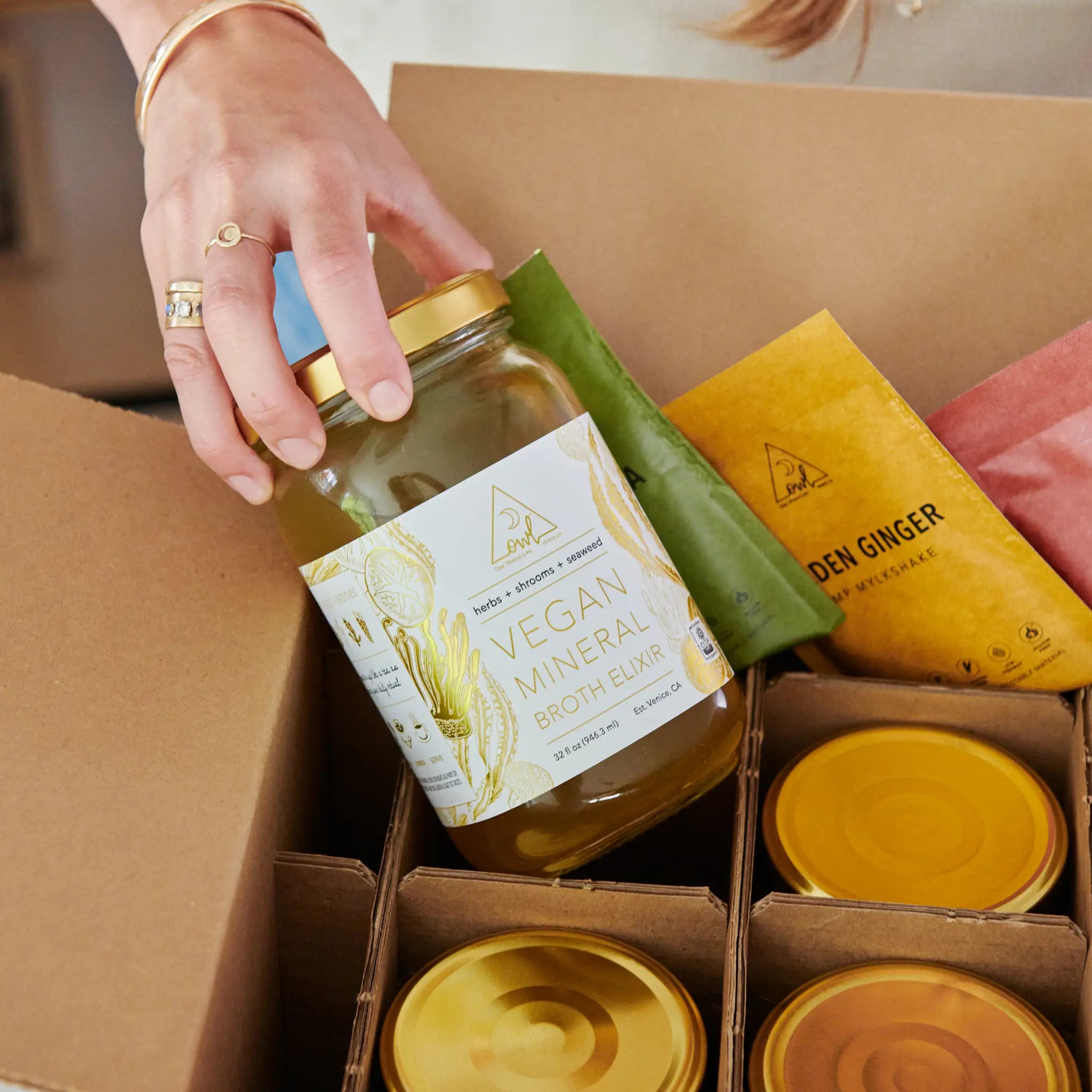 Hand placing a jar of vegan mineral broth elixir into a box with organic cleanse products and wellness drink packets. The OWL Reset - Organic Cleanse Program with 6 Broth Elixirs, 6 Mylkshake Packets (2 Magic Matcha, 2 Golden Ginger, and 2 Bold Beet)