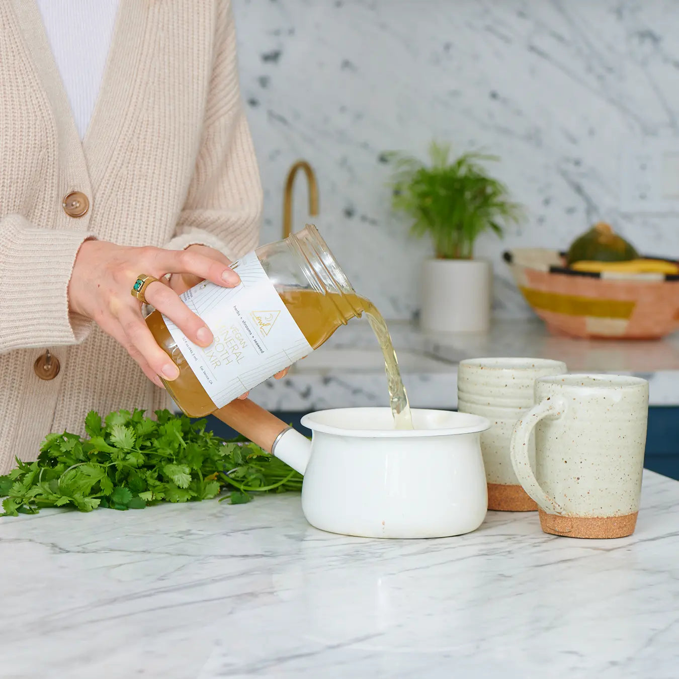 Person pouring liquid from a container into ceramic mugs on a kitchen counter. The OWL Reset - Organic Cleanse Program with 6 Broth Elixirs, 6 Mylkshake Packets (2 Magic Matcha, 2 Golden Ginger, and 2 Bold Beet)