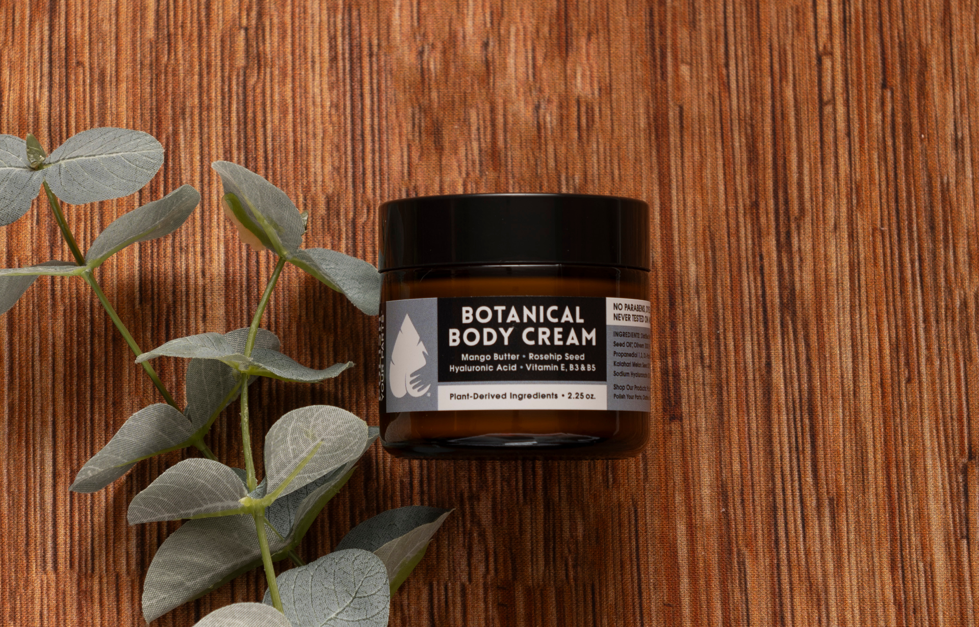 Botanical Body Cream with Mango Butter, Rosehip, Hyaluronic Acid and Vitamin E in an amber jar on a rustic wooden surface with eucalyptus leaves, natural moisturizing skincare, nourishing body cream, plant-derived ingredients, clean beauty, hydrating body care, luxury self-care, Tree of Life Grocers