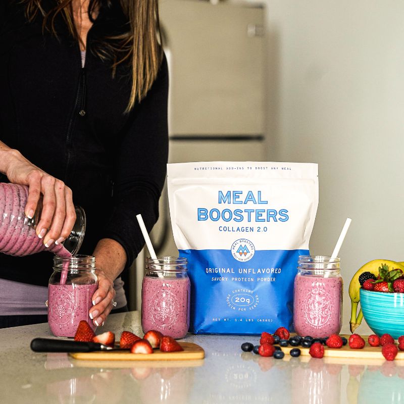Person pouring a pink berry smoothie into mason jars beside **Meal Boosters Original Unflavored Grass-Fed Collagen** in a blue and white resealable pouch, with fresh strawberries, blueberries, bananas, and a bowl of mixed fruit on a bright kitchen countertop.
