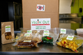 A cardboard box labeled VECIN' OUT with several packaged food items arranged on a counter in front of it, including a brown kraft pouch, a wrapped brown food item, a green salad wrap, and a yellow breakfast-style wrap, in a kitchen setting.