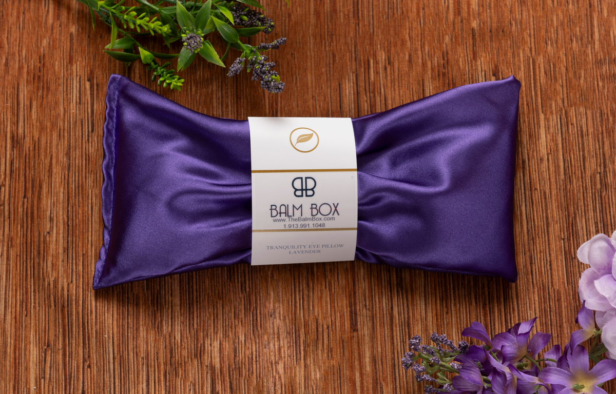 Purple satin lavender tranquility eye pillow with Balm Box label, elegant self-care sleep accessory on rustic wood surrounded by fresh purple flowers, calming aromatherapy relaxation gift, soothing eye pillow for rest, meditation, wellness, stress relief, bedtime, spa and restorative self-care.