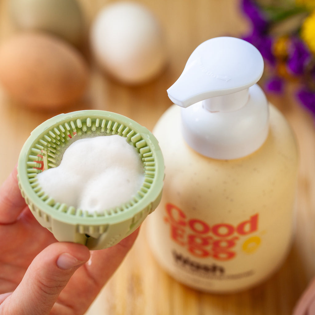 Hand holding the GoodEgg® silicone egg washing brush filled with foaming enzyme egg cleanser beside a GoodEgg® wash bottle, demonstrating an easy, food-safe way to clean fresh backyard, farm, duck, goose, and quail eggs.