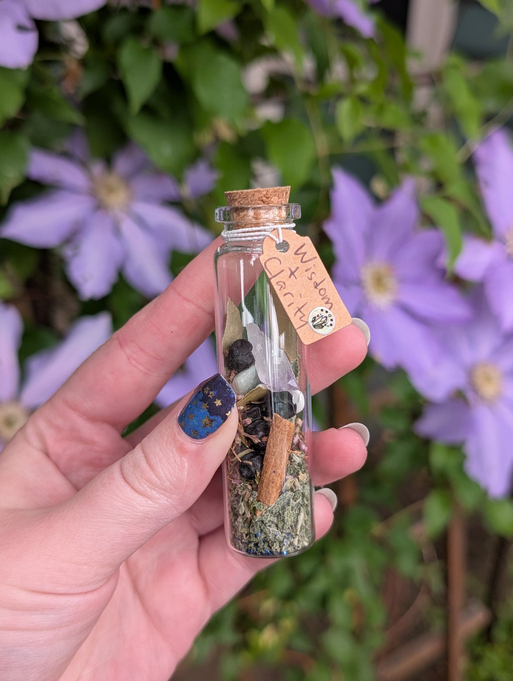 Hand holding a small glass bottle with a cork lid against a background of purple flowers. Wisdom & Clarity Spell Jar