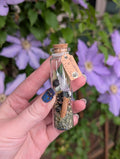 Hand holding a small glass bottle with a cork lid against a background of purple flowers. Wisdom & Clarity Spell Jar