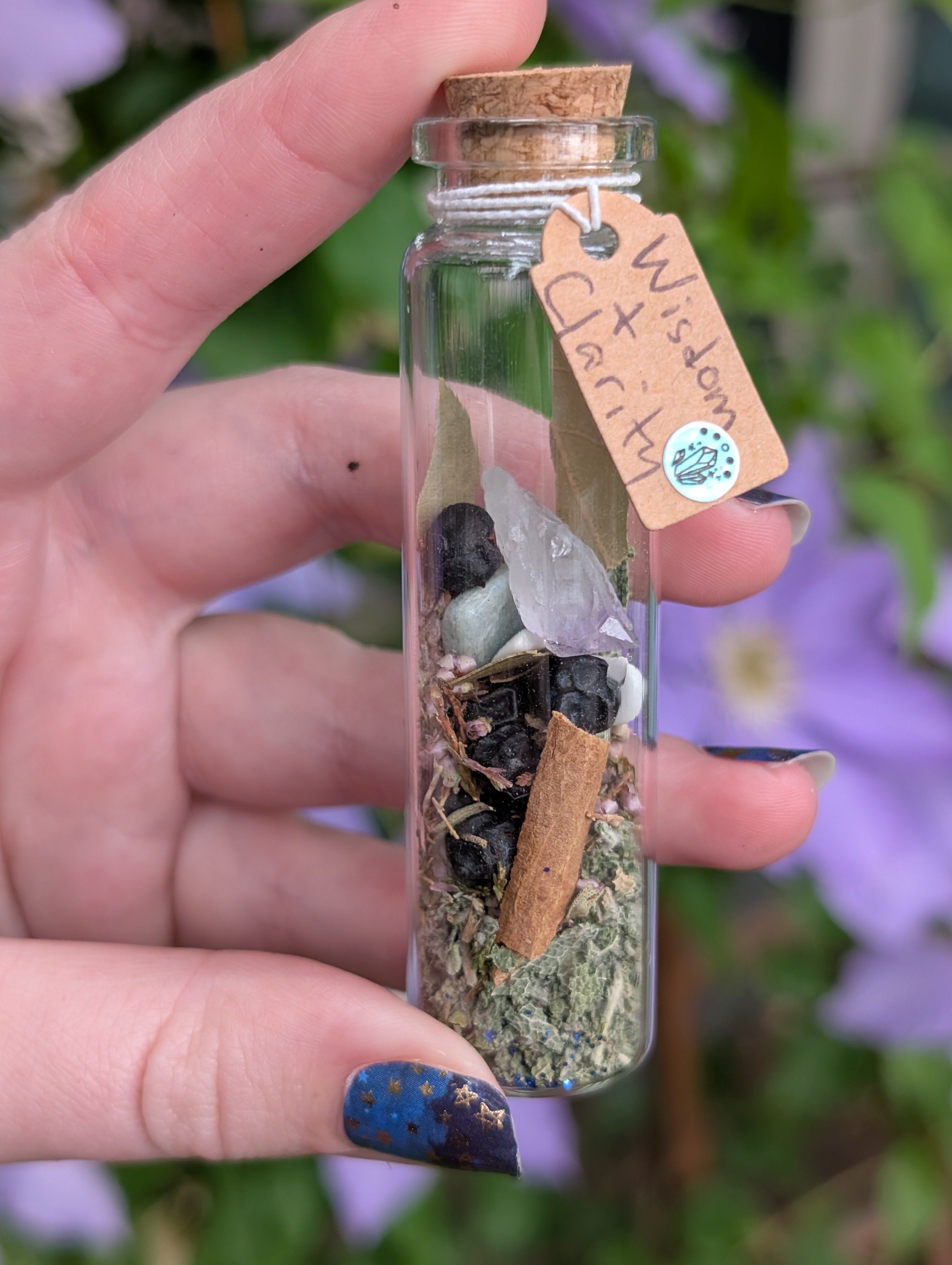 Hand holding a small glass bottle with natural elements and a tag labeled 'Wisdom & Clarity' against a blurred floral background. Wisdom & Clarity Spell Jar