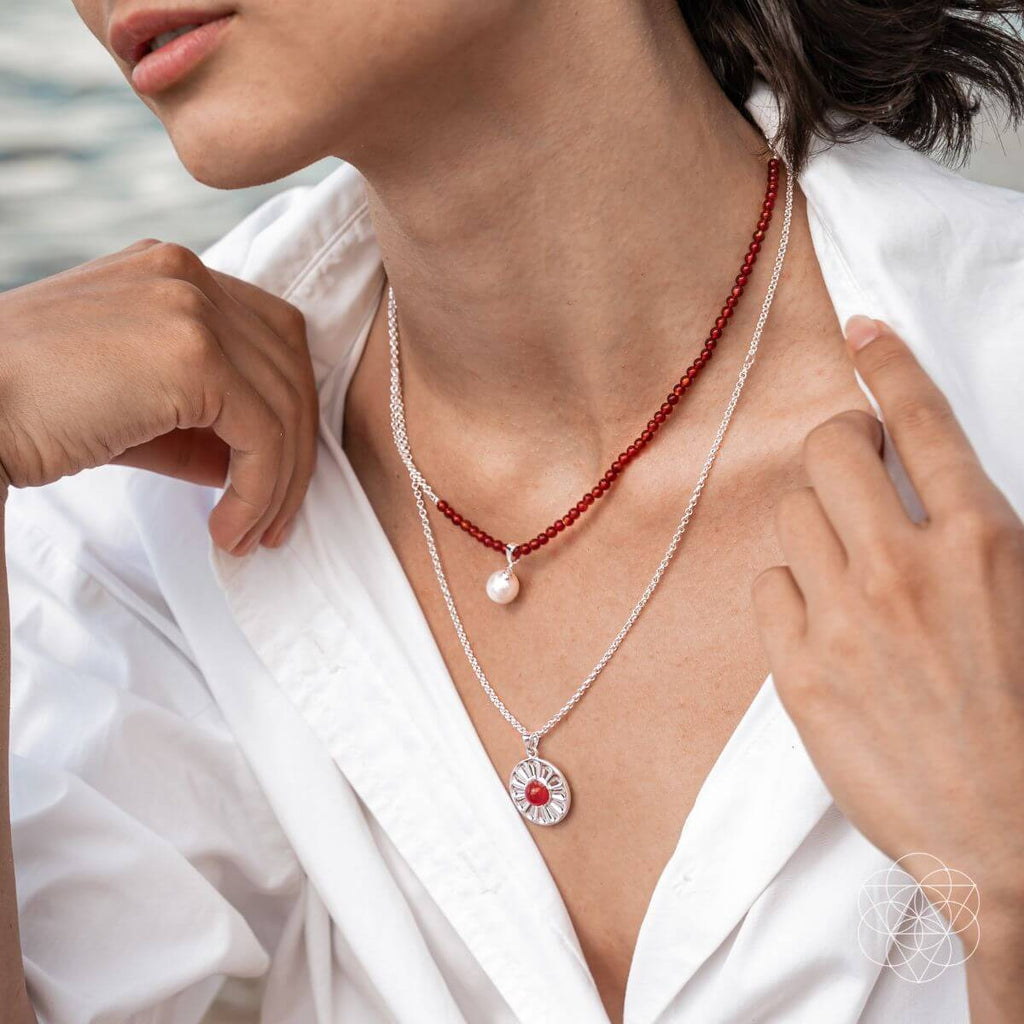 Woman wearing two necklaces with a white shirt, one red and silver, the other pearl and silver. Two layer necklaces with carnelian and freshwater pearl pendants, including a red beaded necklace and a silver pendant with a red carnelian.