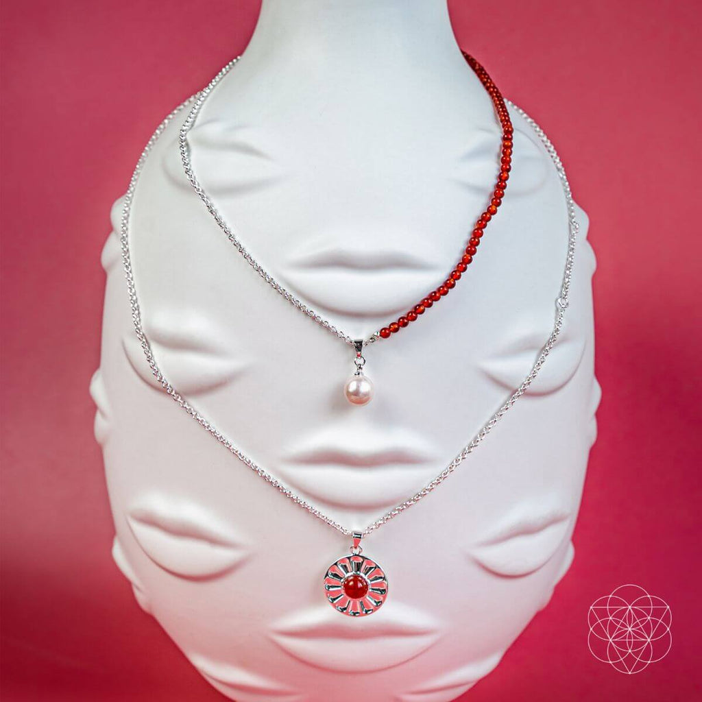 Two necklaces on a white bust against a pink background. Two layer necklaces with carnelian and freshwater pearl pendants, including a red beaded necklace and a silver pendant with a red carnelian.