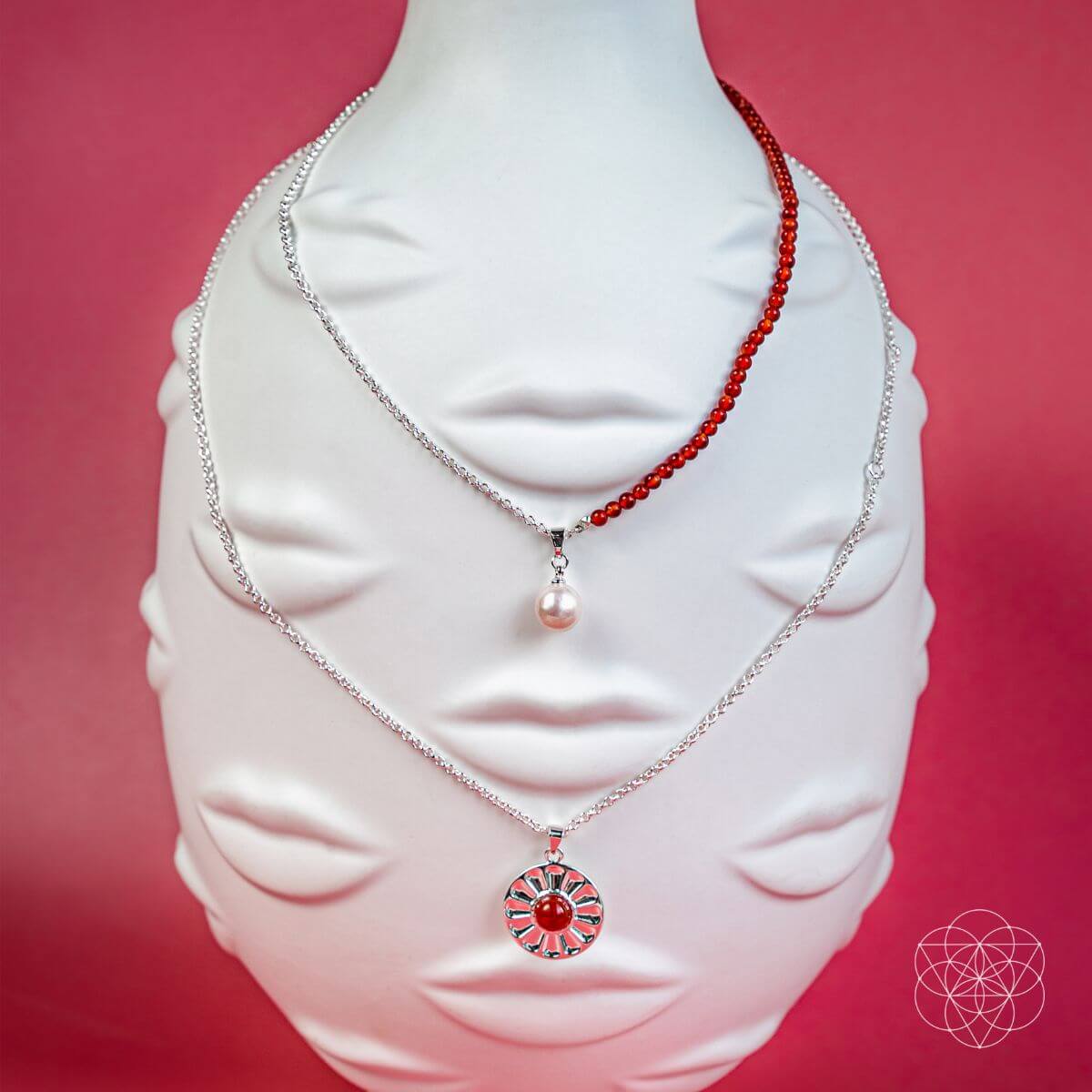 Two necklaces on a white bust against a pink background. Two layer necklaces with carnelian and freshwater pearl pendants, including a red beaded necklace and a silver pendant with a red carnelian.
