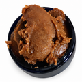 A scoop of 100% Raw Batana Oil: thick, textured brown cosmetic paste in a small dark blue bowl against a plain white background. Stimulates hair follicles and encourages hair growth, nourishes skin, treats dry skin and aids healing scars.