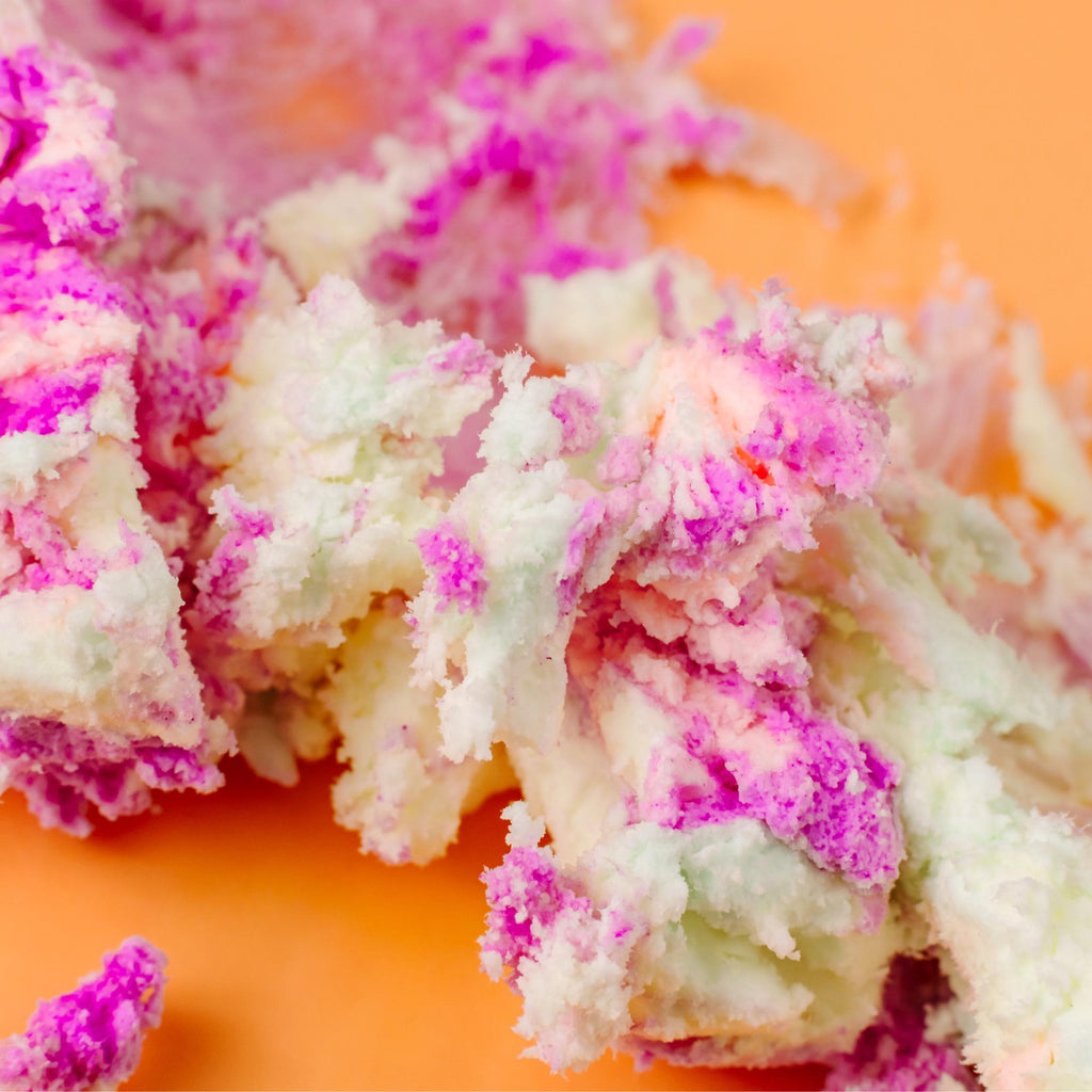 Macro close-up of AMINNAH FRUITY PEBBLEZ Whipped Body Butter featuring its fluffy whipped texture with vibrant purple, pink, peach, and cream swirls on a warm peach background. Rich body butter made with shea butter, mango butter, vitamin E, and essential oils to help moisturize, soften, and nourish dry skin. Clean Beauty, cruelty-free, vegan, made with natural ingredients, female-owned brand, made in the USA, available at Tree of Life Grocers.