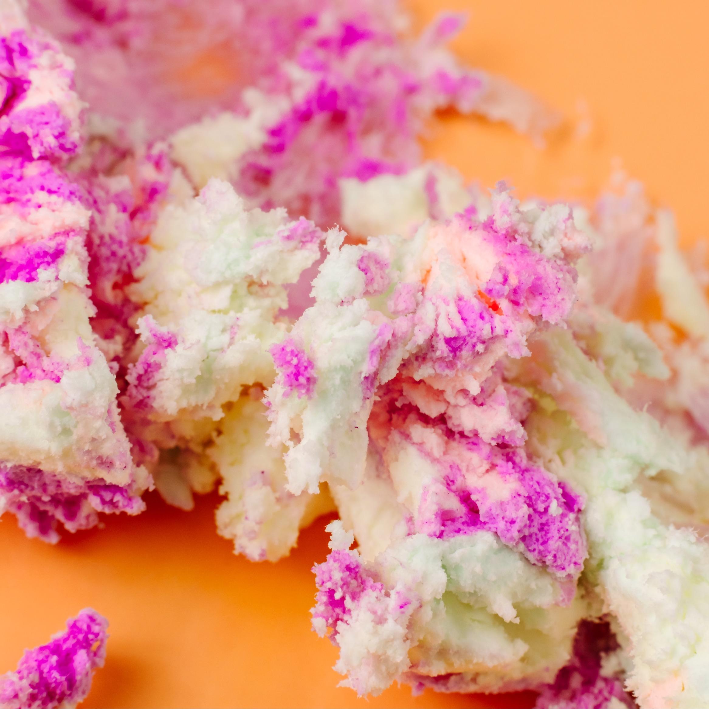 Macro close-up of AMINNAH FRUITY PEBBLEZ Whipped Body Butter featuring its fluffy whipped texture with vibrant purple, pink, peach, and cream swirls on a warm peach background. Rich body butter made with shea butter, mango butter, vitamin E, and essential oils to help moisturize, soften, and nourish dry skin. Clean Beauty, cruelty-free, vegan, made with natural ingredients, female-owned brand, made in the USA, available at Tree of Life Grocers.