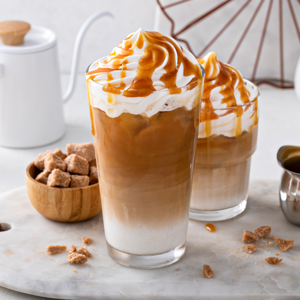 Two tall glasses of iced coffee topped with whipped cream and caramel sauce, styled on a marble surface with sugar cubes and a small white pitcher in the background.