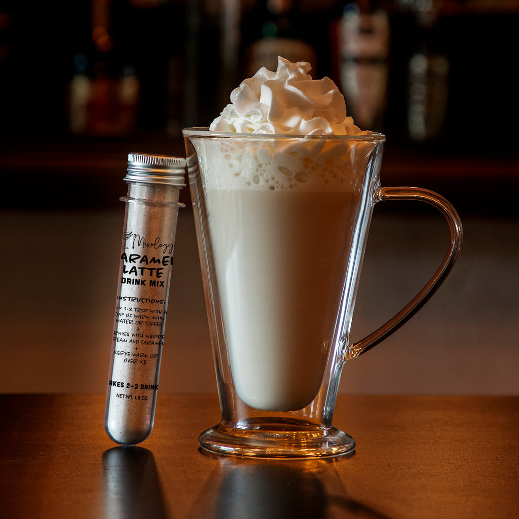 Creamy caramel latte served in a clear glass mug with whipped cream, displayed beside a tube of caramel latte drink mix on a wooden table against a warm, blurred café background.