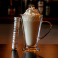Creamy caramel latte served in a clear glass mug with whipped cream, displayed beside a tube of caramel latte drink mix on a wooden table against a warm, blurred café background.