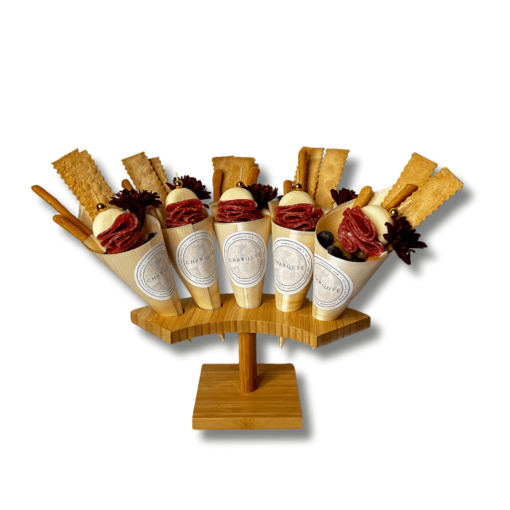 Gourmet charcuterie cones filled with artisan salami, fresh mozzarella, crackers, breadsticks, olives, and decorative floral accents displayed on a wooden serving stand against a white background.