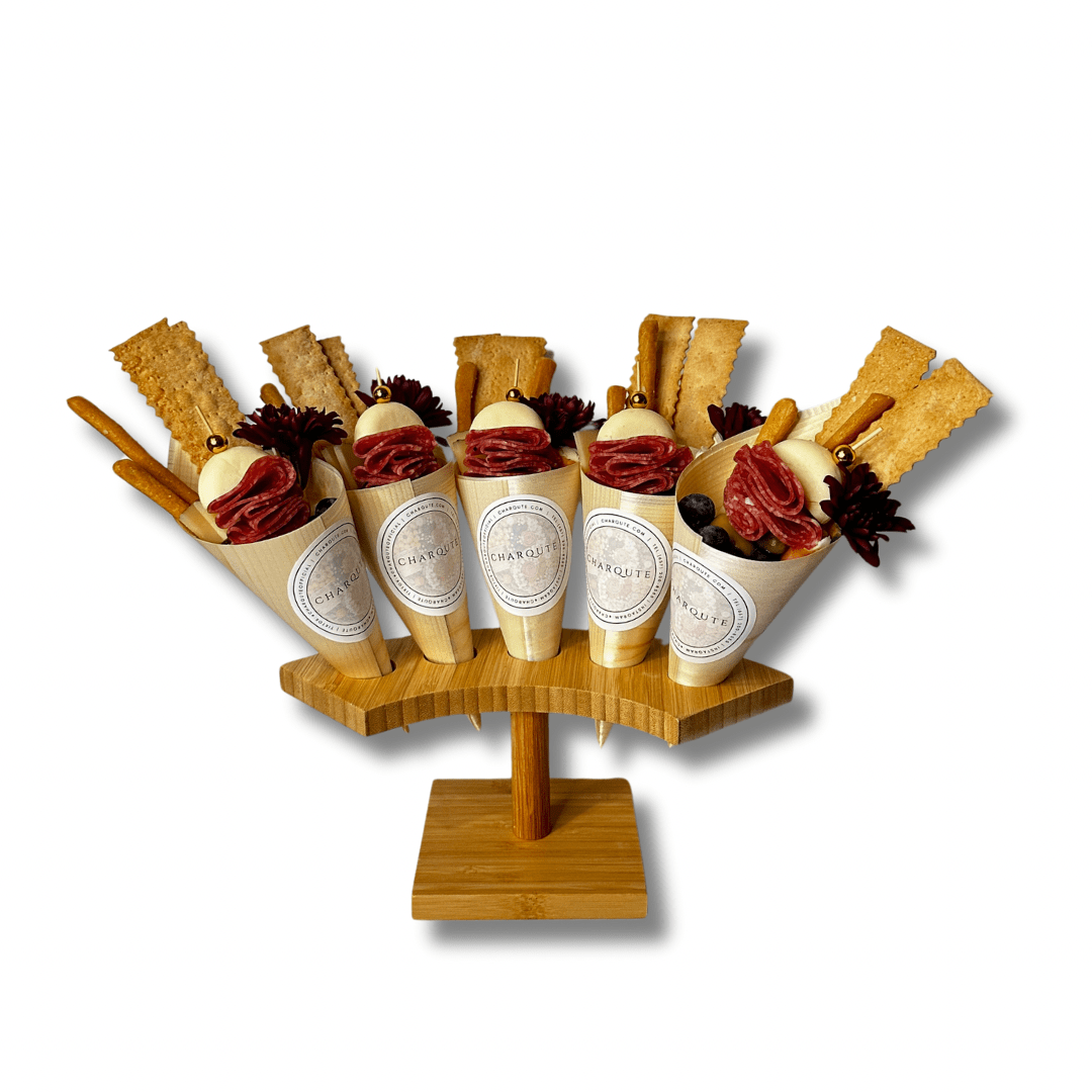 Gourmet charcuterie cones filled with artisan salami, fresh mozzarella, crackers, breadsticks, olives, and decorative floral accents displayed on a wooden serving stand against a white background.