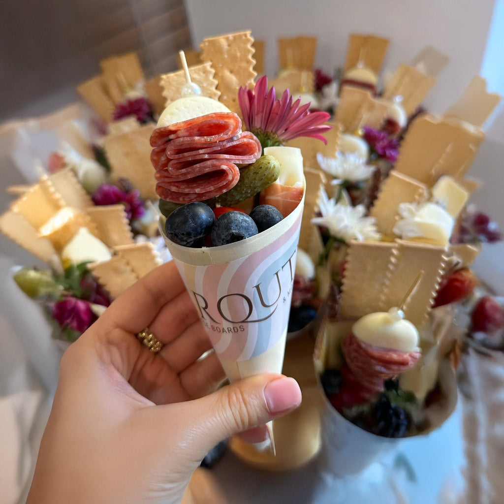 Hand holding a gourmet charcuterie cone filled with salami, mozzarella, crackers, grapes, blueberries, pickle, and fresh flower in front of a display of artisan charcuterie cones.