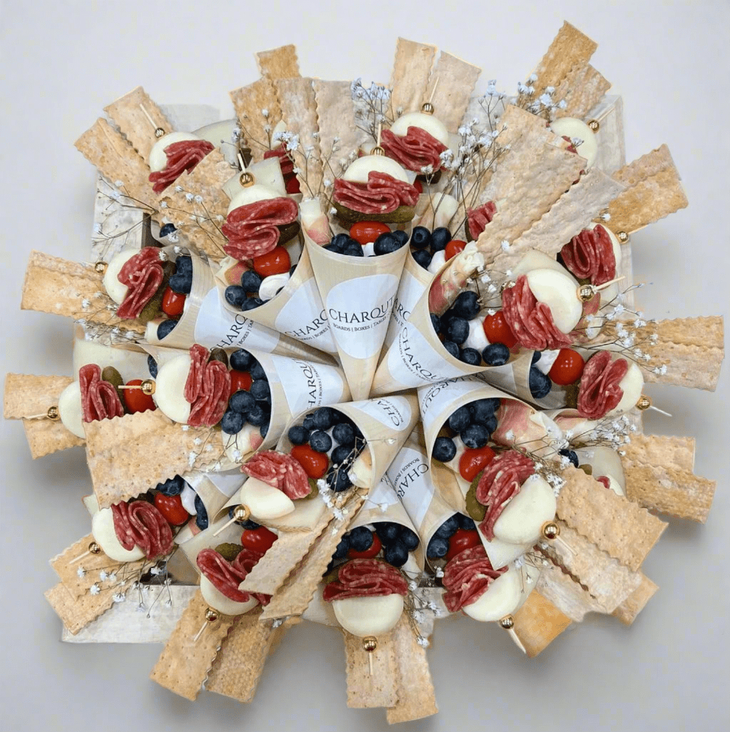 Gourmet charcuterie wreath made with artisan charcuterie cones filled with salami, fresh mozzarella, blueberries, cherry tomatoes, crackers, and decorative floral accents on a white background.