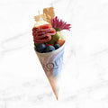Gourmet charcuterie cone with salami, cheese, crackers, olives, grapes, pickle, and fresh flower on a white marble background.