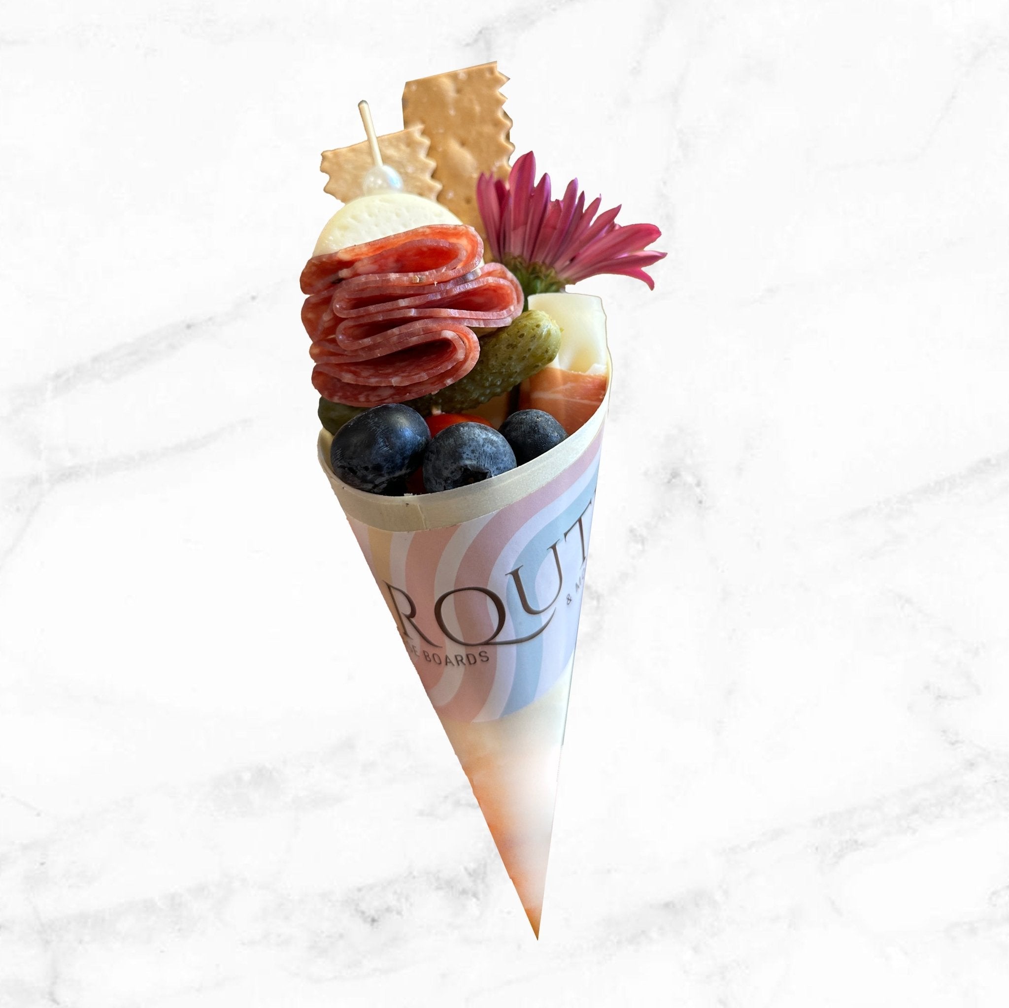 Gourmet charcuterie cone with salami, cheese, crackers, olives, grapes, pickle, and fresh flower on a white marble background.