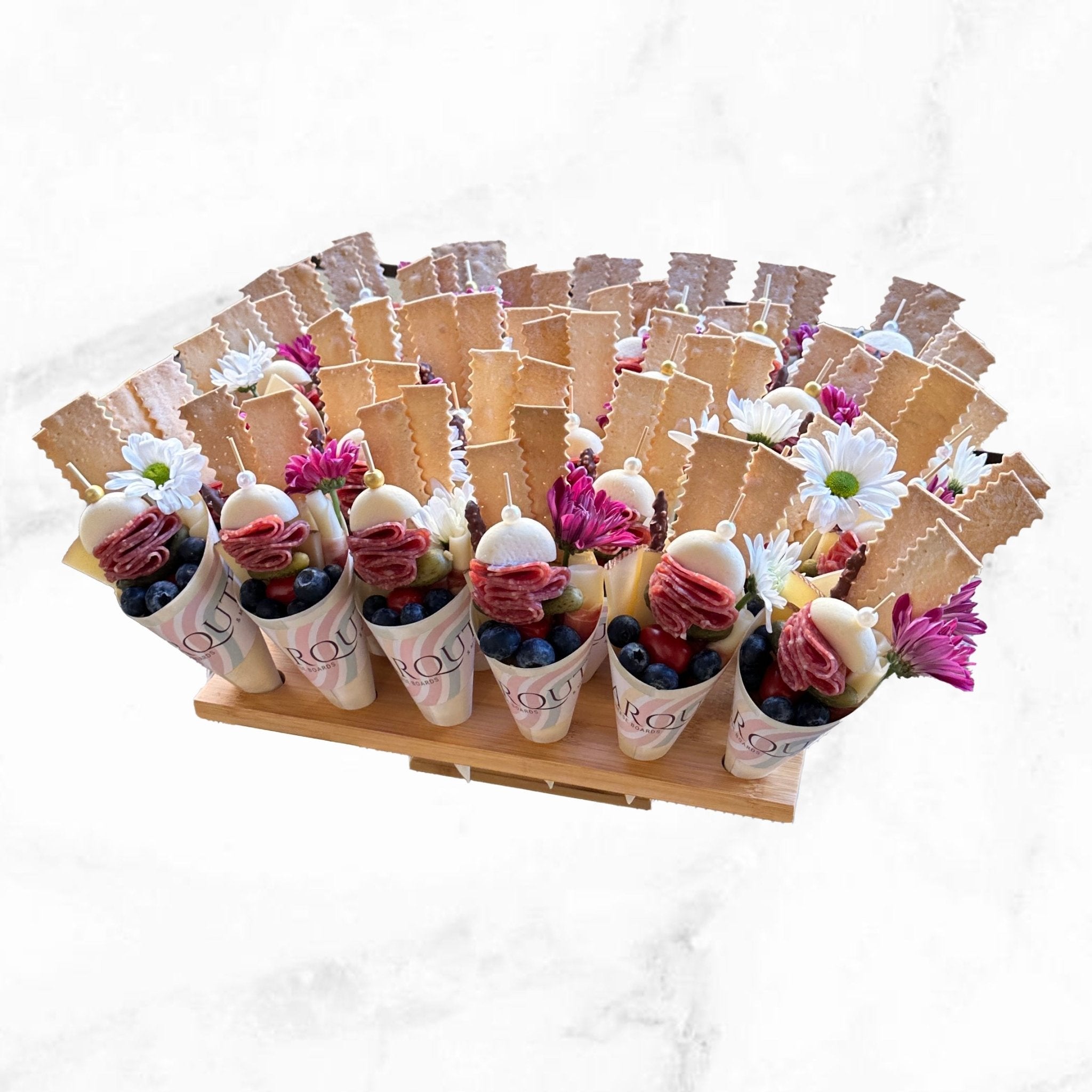 Gourmet charcuterie cones filled with artisan cured meats, cheese, fresh berries, crackers, and edible flowers displayed on a wooden serving stand against a white background.