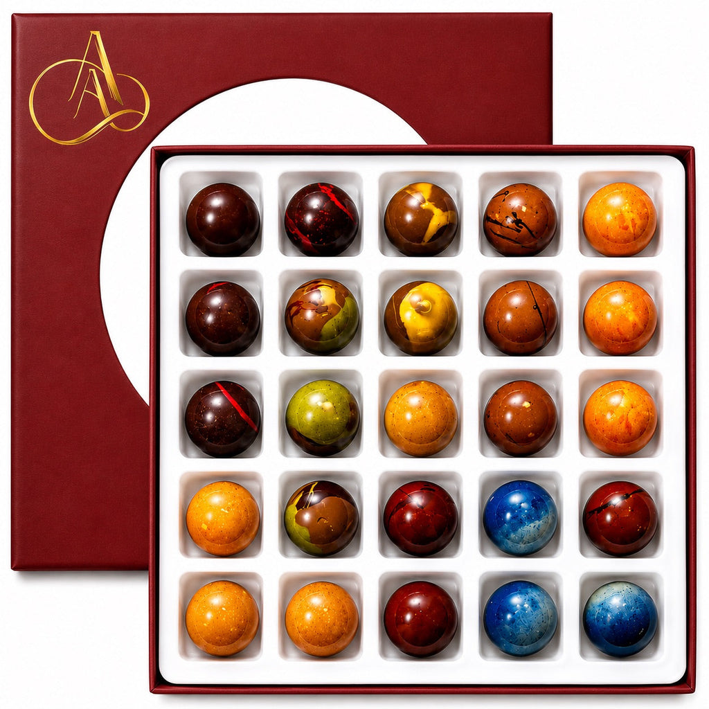 A boxed assortment of marbled gourmet chocolates displayed in a white tray, with the dark red lid partially visible behind it. The chocolates show a variety of colors including orange, brown, red, yellow, green, and blue, and the box features a gold monogram logo.