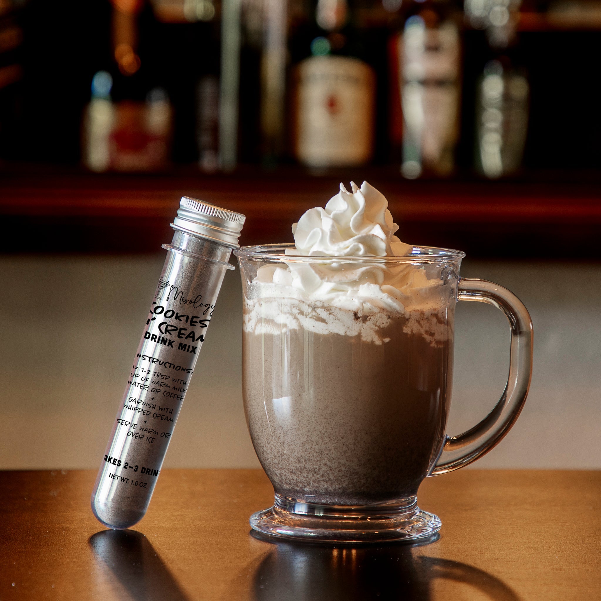 Cookies & Cream hot chocolate mix in a glass mug topped with whipped cream beside a tube of gourmet drink mix on a wooden counter.