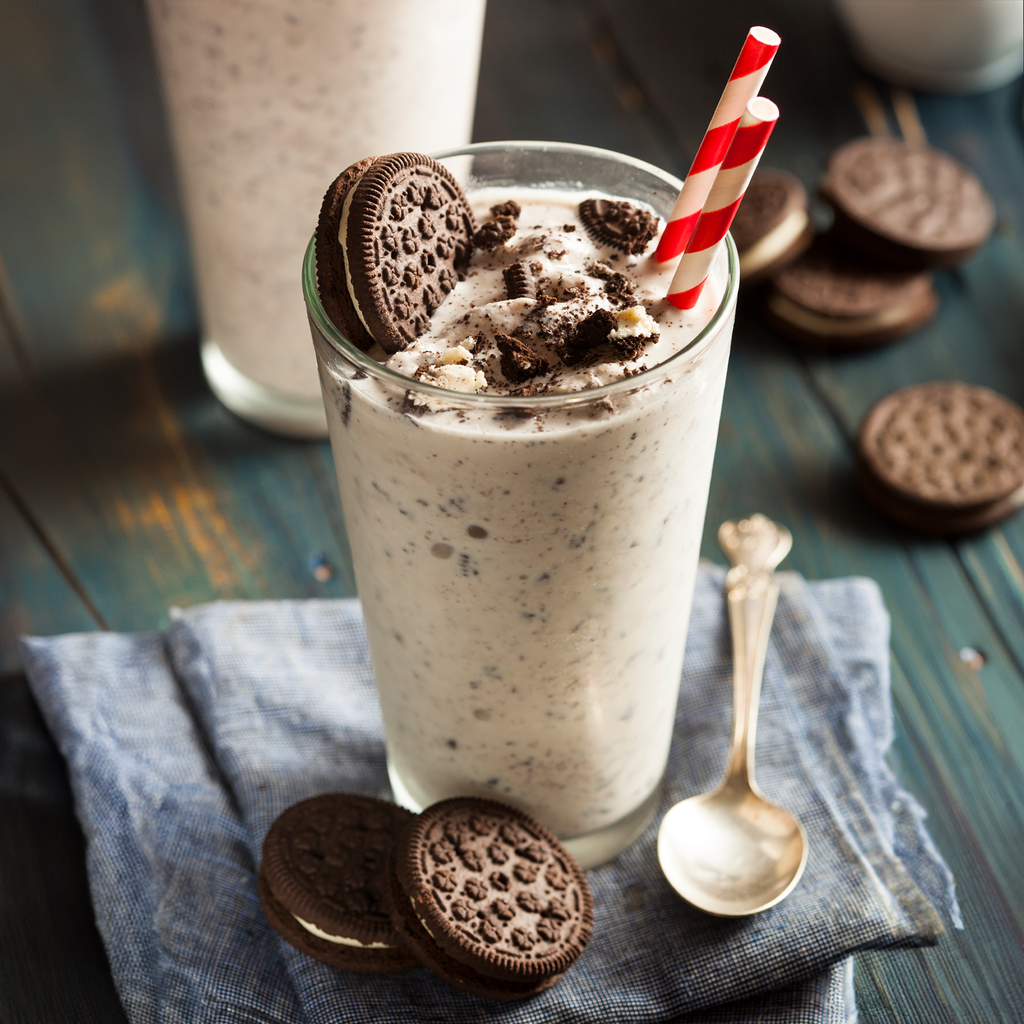 A tall clear glass filled with a creamy cookie milkshake topped with crushed cookie pieces and a red-and-white striped straw, set on a blue cloth with Oreo cookies and a spoon on a rustic dark wooden table.