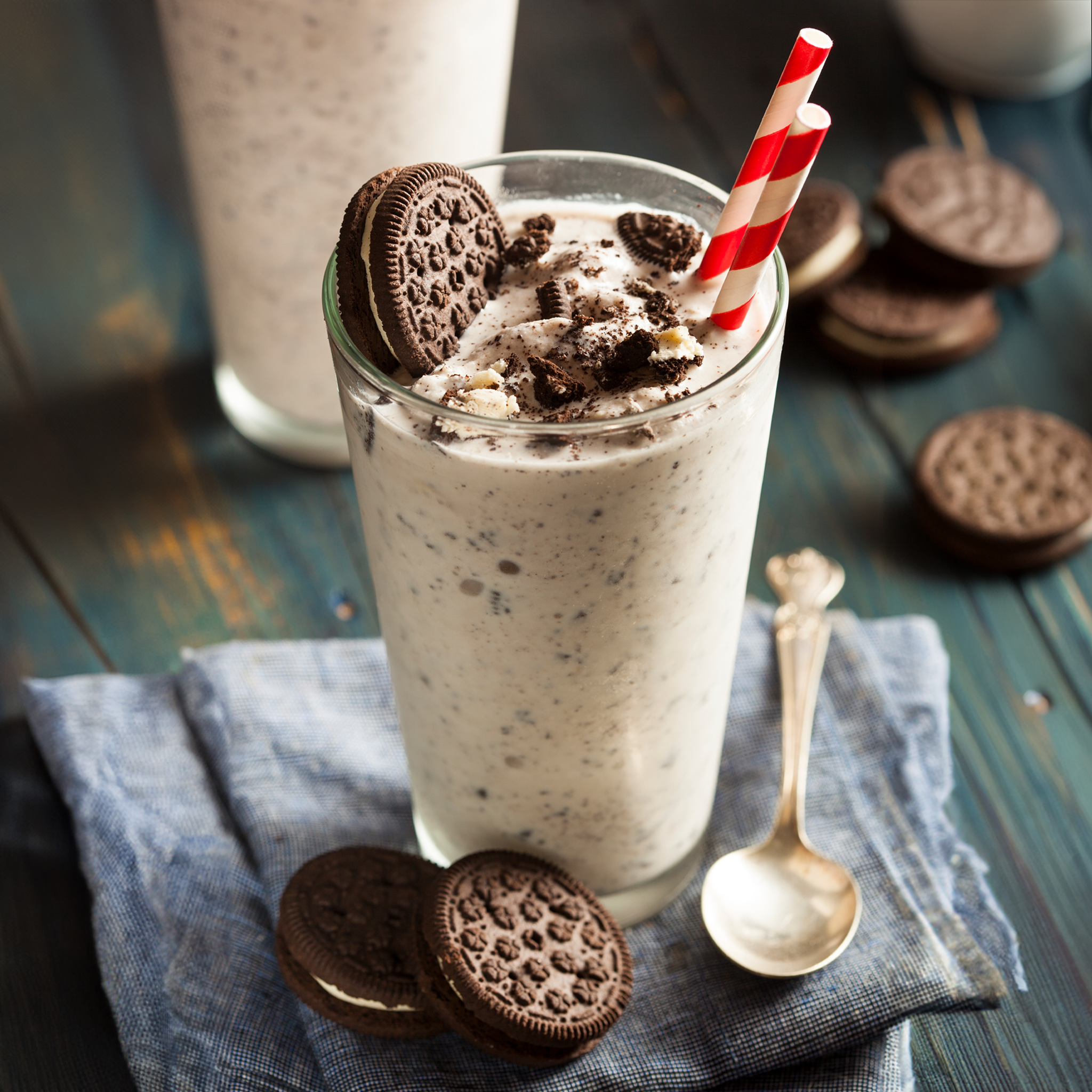 A tall clear glass filled with a creamy cookie milkshake topped with crushed cookie pieces and a red-and-white striped straw, set on a blue cloth with Oreo cookies and a spoon on a rustic dark wooden table.