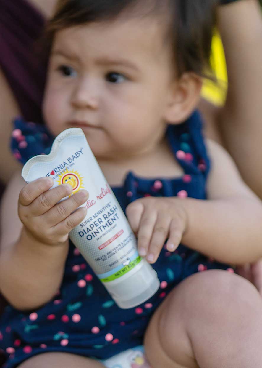 Child holding a bottle of diaper rash ointment with a visible brand logo. California Baby® (No Fragrance) Super Sensitive™ Diaper Rash Ointment