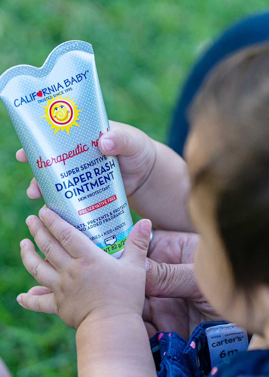 Person holding a California Baby diaper rash ointment tube with a child in the background. California Baby® (No Fragrance) Super Sensitive™ Diaper Rash Ointment