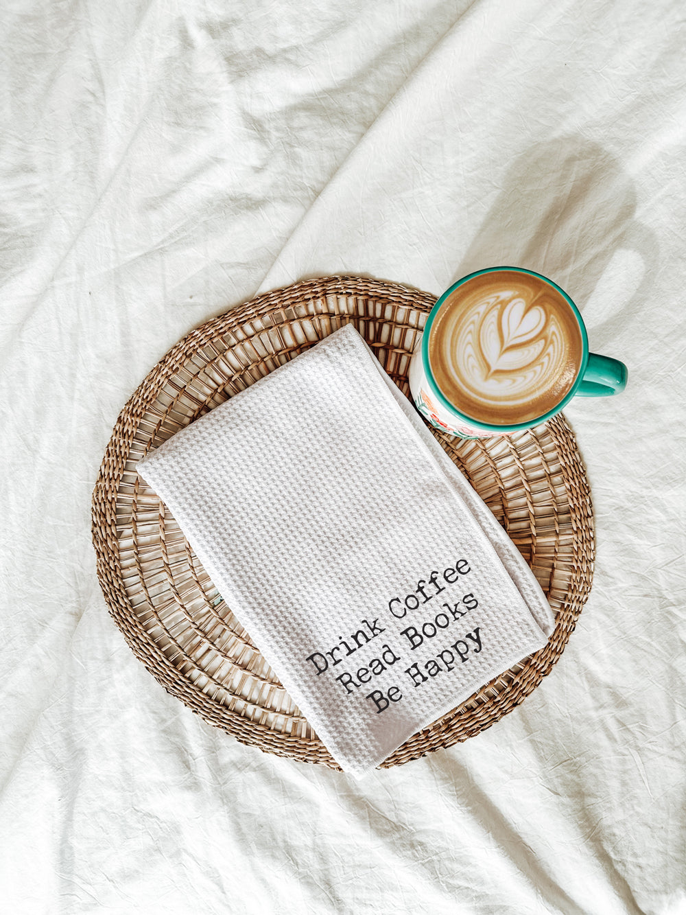 White waffle-weave tea towel embroidered with “Drink Coffee Read Books Be Happy,” styled on a woven placemat beside a latte with leaf art on white fabric.