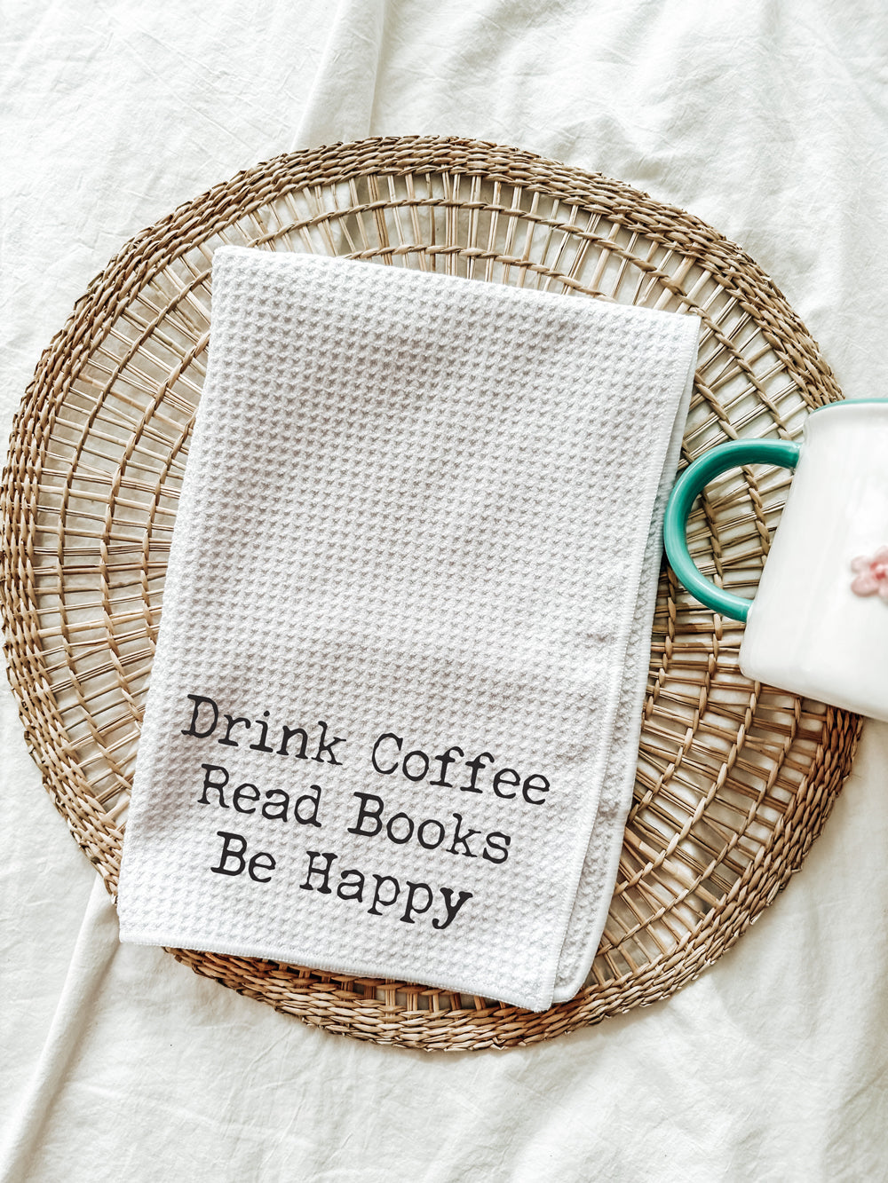 White waffle-weave tea towel embroidered with “Drink Coffee Read Books Be Happy,” styled on a woven placemat beside a white coffee mug.