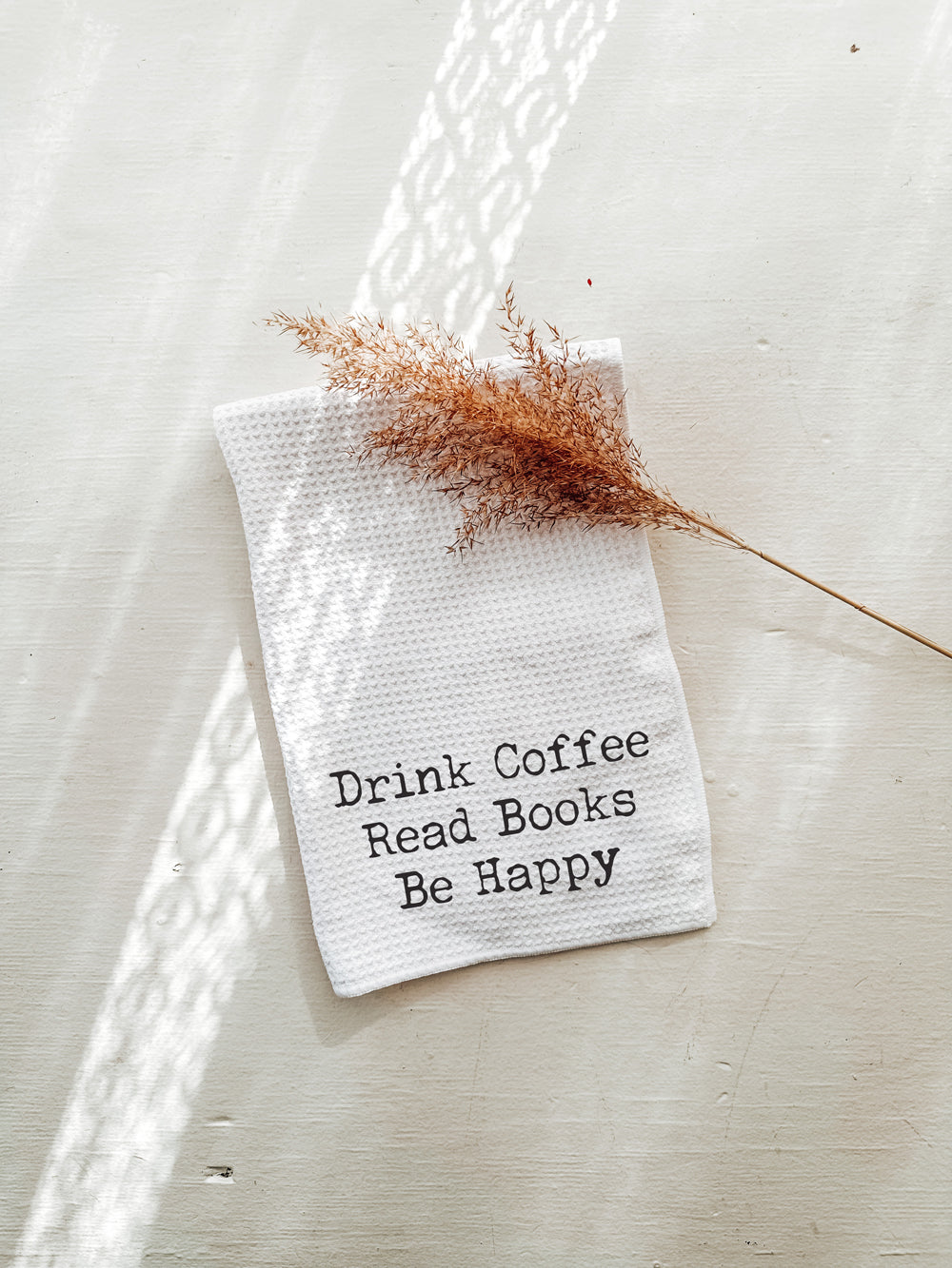 White waffle weave tea towel embroidered with “Drink Coffee Read Books Be Happy,” styled with dried botanicals on a neutral beige background.