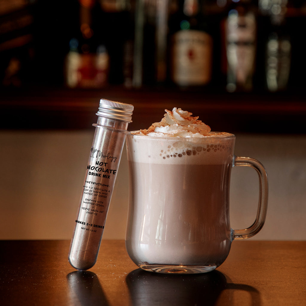 A glass mug filled with a creamy light brown hot chocolate beverage topped with whipped cream sits on a wooden bar counter next to a silver metal tube labeled 'Melted Chocolate Cream'. The background shows a blurred bar shelf with bottles.