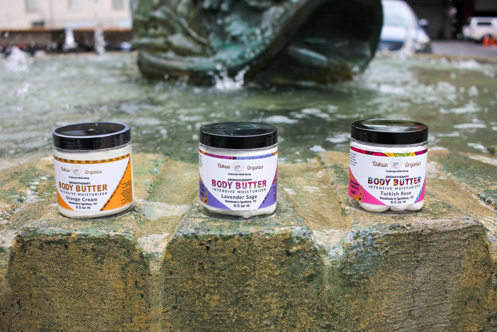 Oshun Organics Aromatherapy Body Butter Intensive Moisturizer in Orange Cream, Lavender Sage and Turkish Rose scents, three jars of rich moisturizing body butter displayed on a stone fountain ledge for soft, smooth, hydrated skin