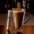 Glass mug filled with a rich, creamy Irish cream–style coffee drink beside a clear tube of Irish Cream Drink Mix on a wooden table with a softly blurred café bar background.