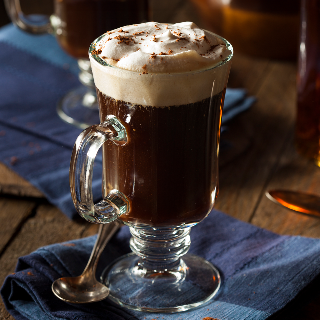 Glass mug of rich Irish cream–style hot drink topped with fluffy whipped cream and cocoa, served on a rustic wooden table with a blue linen napkin and spoon.