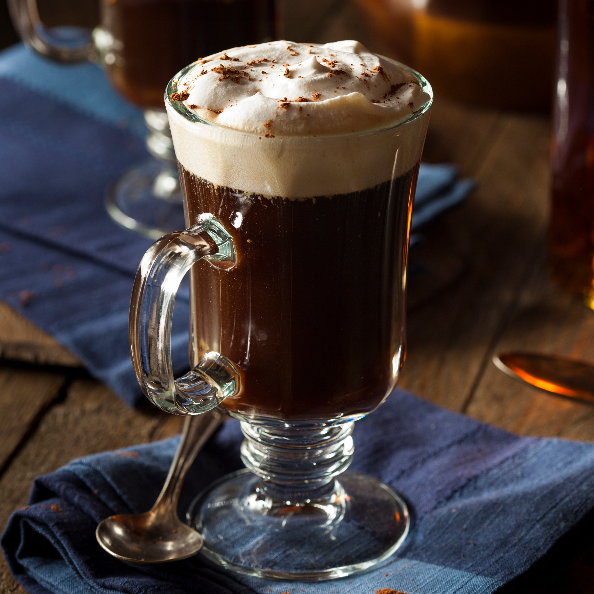 Glass mug of rich Irish cream–style hot drink topped with fluffy whipped cream and cocoa, served on a rustic wooden table with a blue linen napkin and spoon.