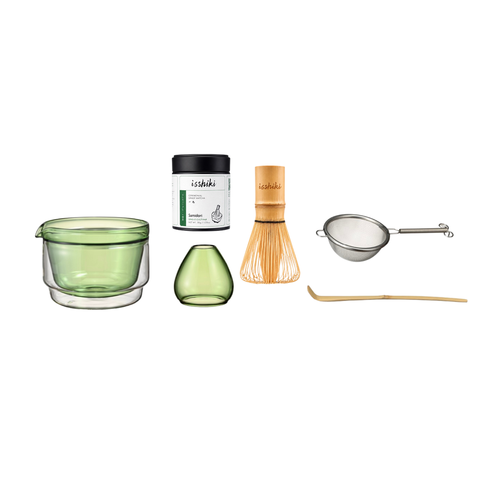 Set of tea-making tools including a green glass cup, small green vase, black container, wooden whisk, metal sieve, and wooden spoon on a white background.