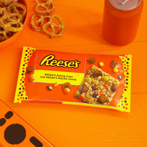 Reese's baking cups packaging on an orange surface with pretzels and a can.