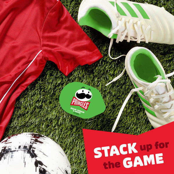Red sports jersey, green and white sneakers, soccer ball, and Pringles chip bag on grass with 'Stack up for the Game' text.