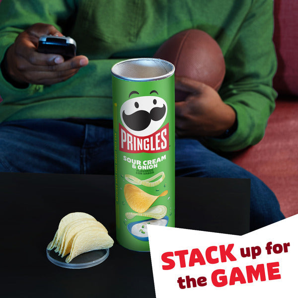 Pringles Sour Cream & Onion potato crisps can on a table with stacked chips in front, a person holding a football and TV remote in the background, and "Stack up for the Game" promotional sign.