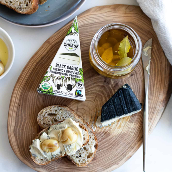 Black garlic dairy-free cashew cheese served on artisan bread with pickled garlic on a rustic wooden board, featuring organic vegan cheese wedge packaging.