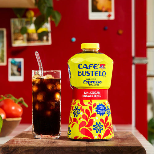 Cafe Bustelo coffee bottle with colorful design next to a glass of iced coffee on a wooden surface. Café Bustelo Unsweetened Espresso-Style Iced Coffee