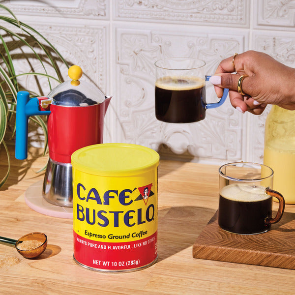 A vibrant display of **Café Bustelo Espresso Style Dark Roast Ground Coffee** in its iconic yellow can beside a colorful stovetop espresso maker, with two freshly brewed glasses of rich black coffee on a wooden countertop, creating a warm and inviting coffeehouse-inspired kitchen scene.